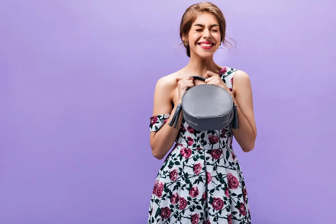 Mädchen in bester Laune hält eine graue Handtasche. Glückliche, charmante junge Frau im Kleid mit Blumenmuster posiert mit einer kleinen, trendigen Tasche vor isoliertem Hintergrund.